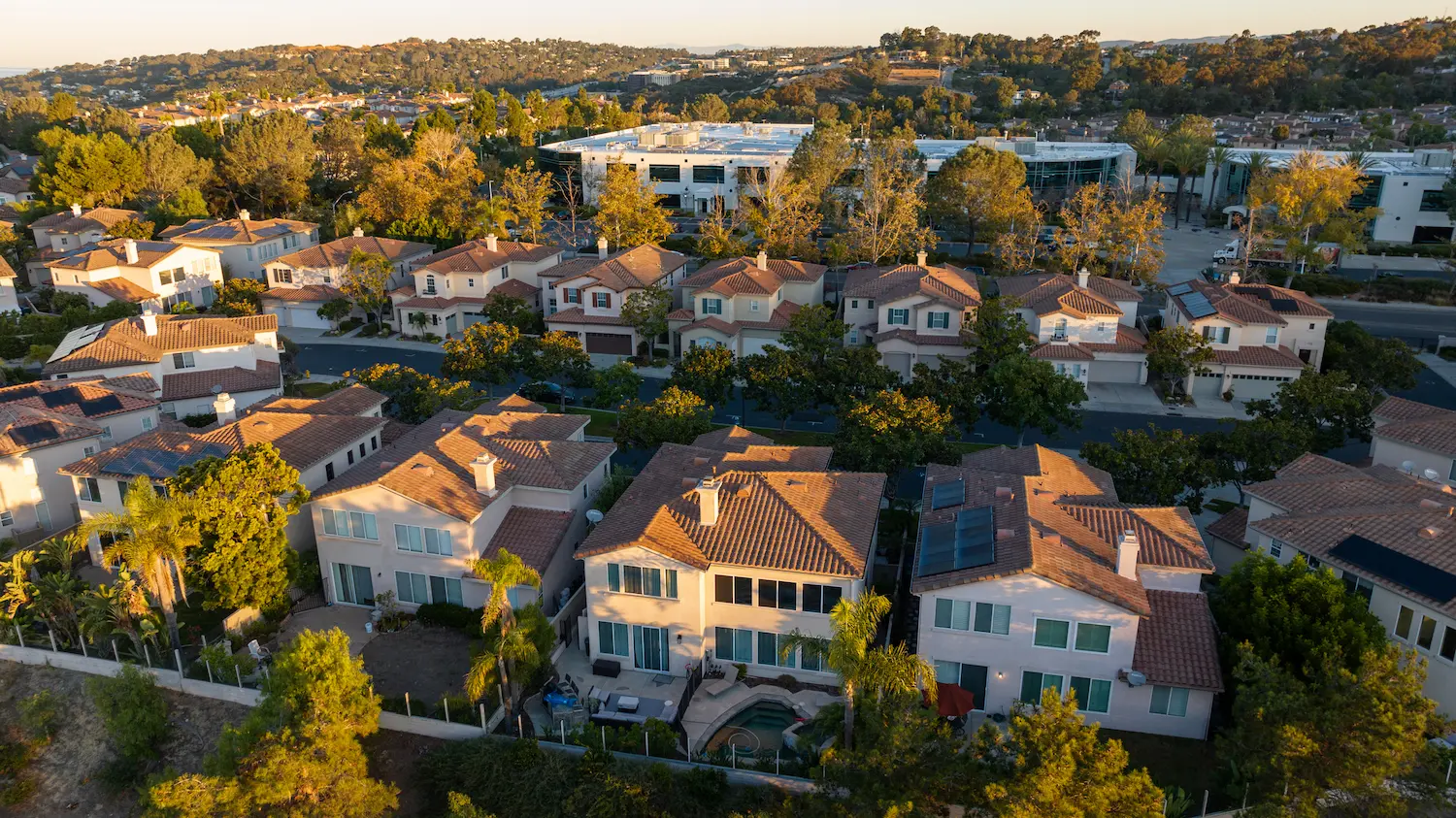 Golden hour aerial view of suburban homes with tile roofs and solar panels near a commercial office park in a lush, hilly community.